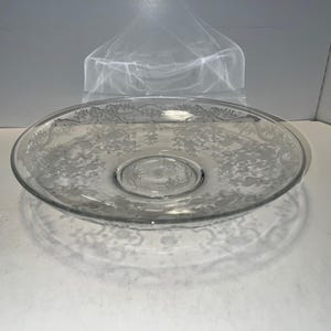 Puede incluir: Plato de servir de vidrio transparente con un diseño floral y de follaje grabado alrededor del borde y el centro. El plato es redondo y se encuentra sobre una superficie blanca. El diseño grabado es visible.