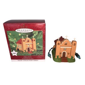May include: A Hallmark Keepsake Ornament of an adobe church with a red roof, crosses, and a green base. The ornament is tan with a brown roof and has a power cord. The box reads "Adobe Church Candlelight Services".