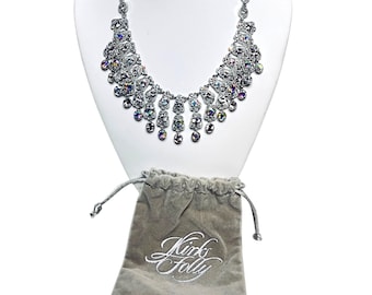 May include: A silver necklace with iridescent crystal embellishments is displayed on a white bust. The necklace has a bib design with dangling teardrop crystals. A grey velvet pouch with the embroidered text "Kirks Folly" is at the base.