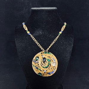 May include: A gold chain necklace with a large, round pendant. The pendant is made of gold and features a swirling design with green and blue gemstones.