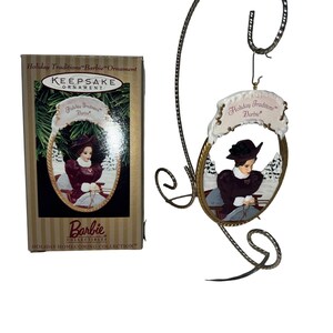 May include: A Barbie Holiday Homecoming Collection ornament. The oval-shaped ornament features a woman in a burgundy dress and black hat, set against a snowy backdrop. The ornament is displayed on a decorative metal stand, with the original box visible.