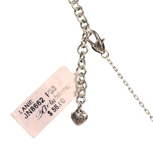 May include: Close-up of a silver necklace with a lobster clasp and a heart-shaped charm. The necklace has a chain link design. A white tag with the text "LANIE JN8662 F33 Brighton" is attached to the necklace.