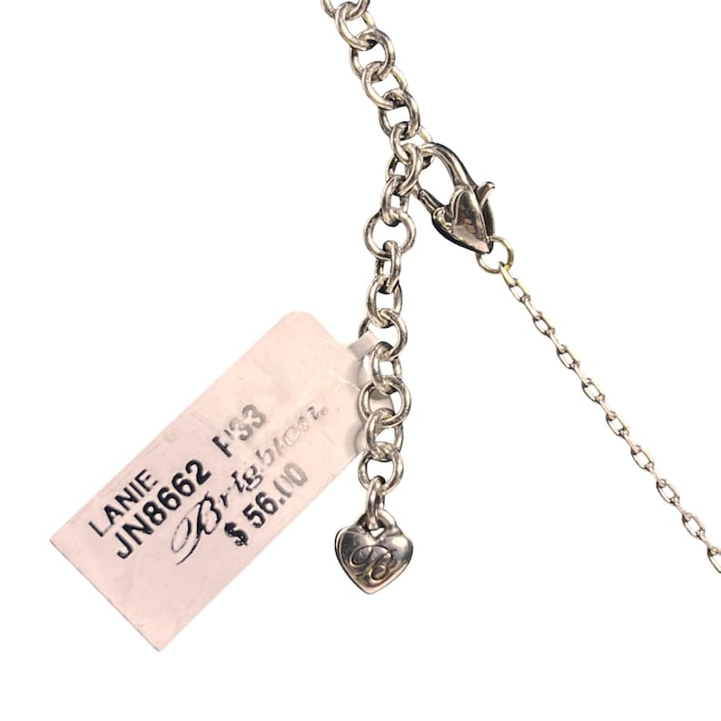 May include: Close-up of a silver necklace with a lobster clasp and a heart-shaped charm. The necklace has a chain link design. A white tag with the text "LANIE JN8662 F33 Brighton" is attached to the necklace.
