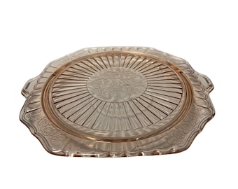 Puede incluir: Un plato para pastel de vidrio rosa con un borde festoneado y un diseño floral en el centro. El plato tiene un patrón de rayos de sol en relieve alrededor del borde.