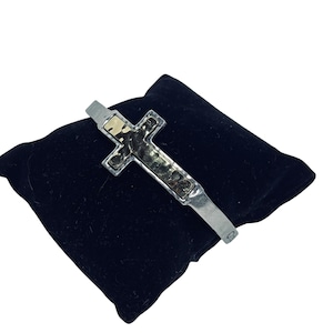 May include: Silver-tone cross bracelet with a textured gold-tone center, resting on a black velvet surface. The cross design is the focal point, showcasing a blend of metallic finishes and intricate detailing.