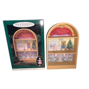 May include: A Keepsake Ornament depicting a miniature stage scene from "The Nutcracker Ballet." The ornament is a wooden shelf design with a ballerina figurine, a Christmas tree, and stage curtains. The box is green with the ornament's details.