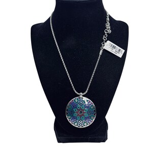 May include: Silver necklace with a circular pendant. The pendant has a detailed, colourful design in shades of blue, green, and red. The necklace is displayed on a black mannequin.