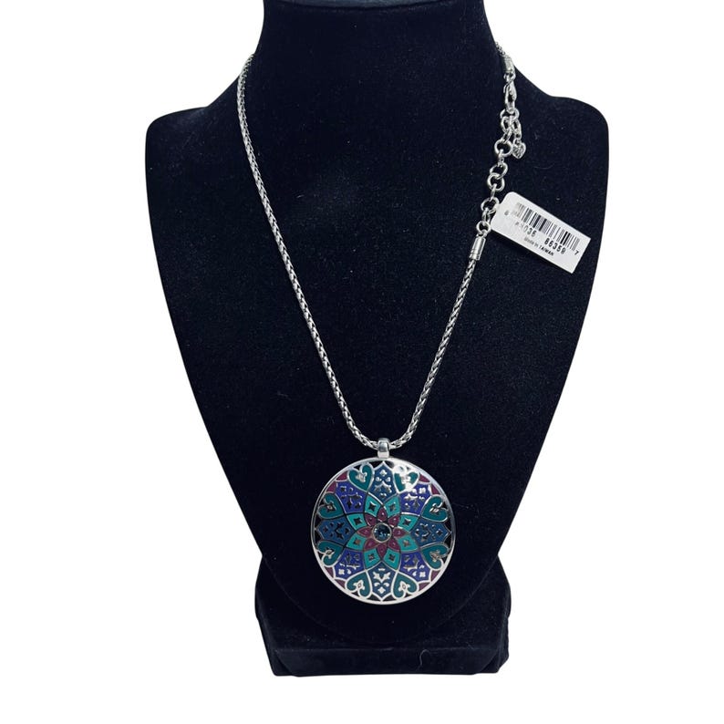 May include: Silver necklace with a circular pendant. The pendant has a detailed, colourful design in shades of blue, green, and red. The necklace is displayed on a black mannequin.