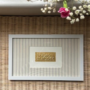 May include: A white-framed artwork with a fabric background of vertical black stripes. A gold plaque displays the coordinates 37.424911, 25.40296E. Decorative flowers are in the background, adding a touch of elegance to the piece.