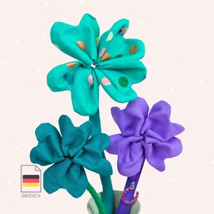 Könnte beinhalten: Ein Strauß Stoffblumen in Türkis- und Lilatönen. Eine Blume hat türkisfarbene Blütenblätter mit goldenen und rosa Tupfen. Die anderen Blumen sind einfarbig türkis und lila. Die Blumen stehen in einer kleinen Vase mit grünem Stiel.