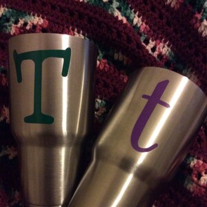 May include: Two stainless steel tumblers with personalized letters. The tumbler on the left has a green "T" and the tumbler on the right has a purple "t".