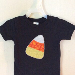 Op de afbeelding: Zwart T-shirt met een vilt applique in de vorm van een snoepmais. De snoepmais is geel, oranje en wit.