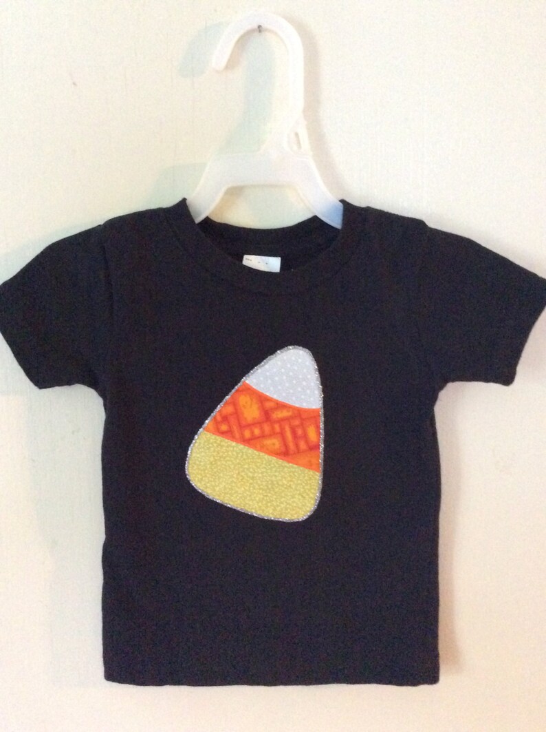 Op de afbeelding: Zwart T-shirt met een vilt applique in de vorm van een snoepmais. De snoepmais is geel, oranje en wit.