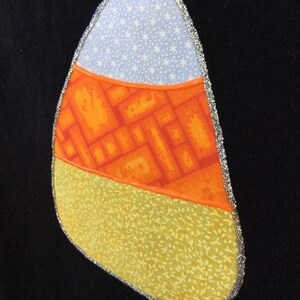 Op de afbeelding: Een patch in de vorm van een snoepmais met een wit, oranje en geel stoffen ontwerp. De patch is omrand met zilveren glitter.