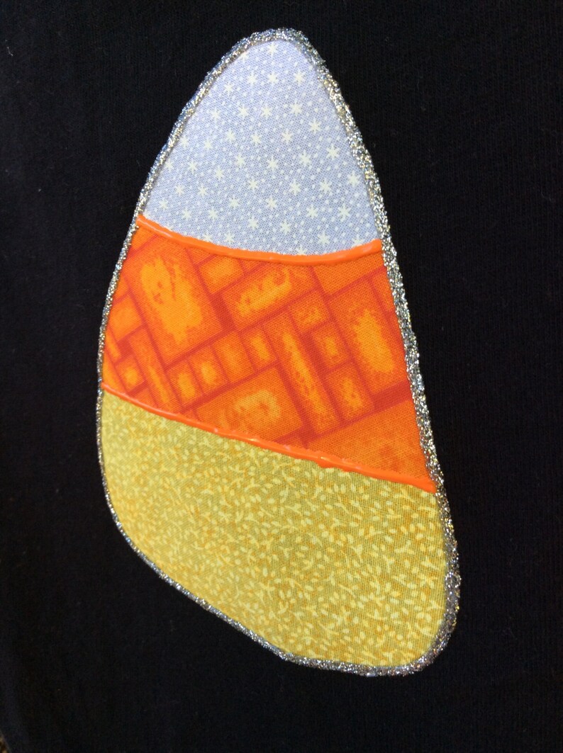 Op de afbeelding: Een patch in de vorm van een snoepmais met een wit, oranje en geel stoffen ontwerp. De patch is omrand met zilveren glitter.