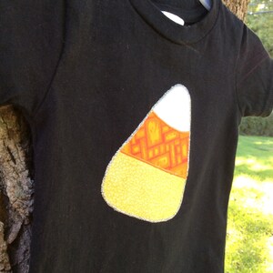 Op de afbeelding: Zwart T-shirt met een vilt applique in de vorm van een snoepmais. De snoepmais is geel, oranje en wit met een zilveren glitterrand.