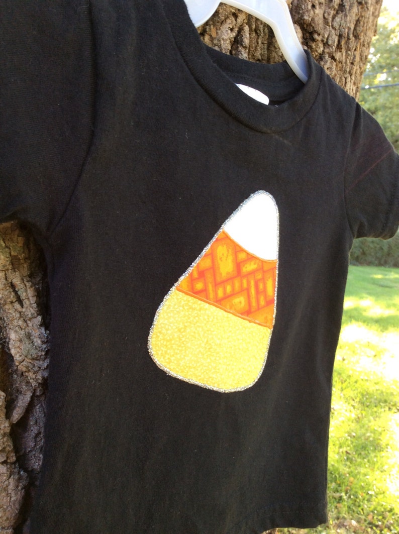 Op de afbeelding: Zwart T-shirt met een vilt applique in de vorm van een snoepmais. De snoepmais is geel, oranje en wit met een zilveren glitterrand.