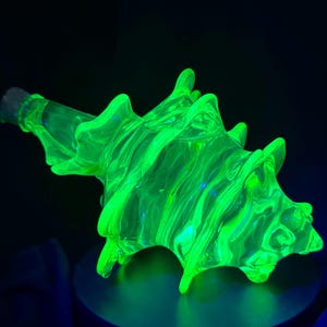 May include: A neon green glass bottle with a unique, spiky design. The bottle has a textured surface and a dark stopper. The vibrant green color glows against a dark background, highlighting its unusual shape.