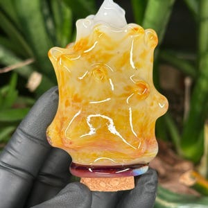May include: A decorative glass bottle stopper featuring an irregular, organic shape with a translucent yellow and orange marbled pattern. The base of the glass section is accented with a dark purple ring, and it is fitted with a natural cork stopper.