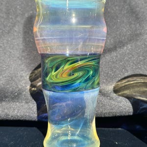 May include: A hand-blown glass with a flared top and a rounded base. The glass features a swirling design in shades of green, blue, and yellow. The upper portion has horizontal bands of color, and the overall effect is iridescent.