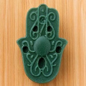 May include: A dark green Hamsa hand-shaped decorative item with detailed, swirling patterns and three small holes. The item is set against a light brown, wood-grain background.