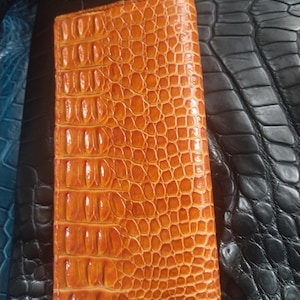 May include: A rectangular, orange wallet with a textured, crocodile-like pattern. The wallet is made of leather and has a glossy finish. The wallet is lying on a black leather surface.