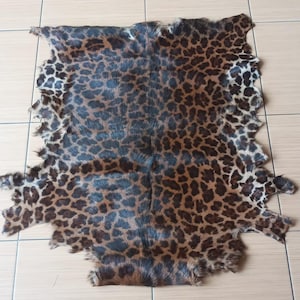 May include: A rectangular animal hide featuring a dense, short-haired coat with a prominent leopard-style pattern. The fur displays a mix of dark brown, black, and tan rosettes and spots across the entire surface, with irregular, jagged edges.