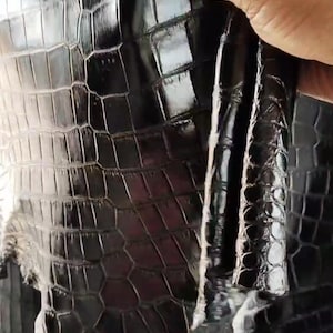 May include: Close-up of black leather with a crocodile skin pattern. The leather has a glossy finish and a textured surface. The image shows the material's details, highlighting its scales and overall texture. The leather is being held.