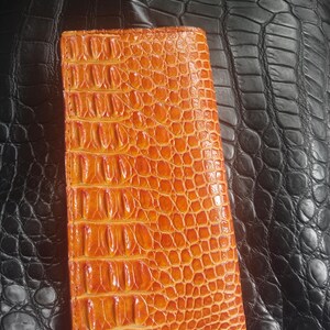 May include: An orange, rectangular wallet with a textured, crocodile-skin pattern. The wallet is made of leather and has a vertical design. The background features a black leather surface.