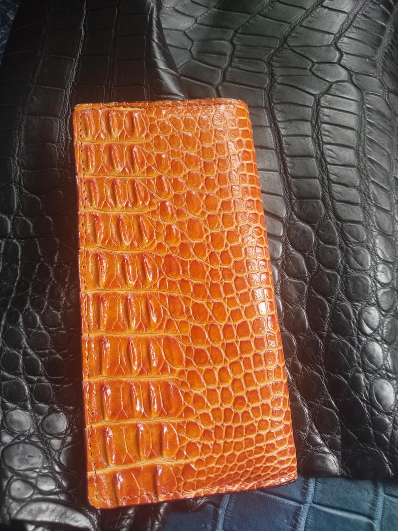 May include: An orange, rectangular wallet with a textured, crocodile-skin pattern. The wallet is made of leather and has a vertical design. The background features a black leather surface.
