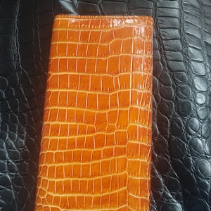 May include: A rectangular, orange leather wallet with a crocodile skin texture. The wallet is closed and appears to be made of high-quality leather. The background is a black, textured surface.