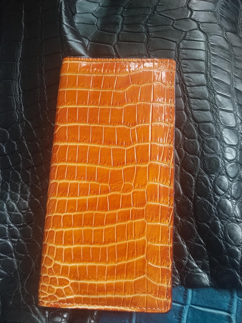 May include: A rectangular, orange leather wallet with a crocodile skin texture. The wallet is closed and appears to be made of high-quality leather. The background is a black, textured surface.