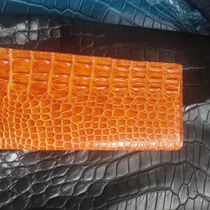 May include: An orange leather wallet with a textured crocodile pattern. The wallet is rectangular and lies on a surface of black and blue leather with a similar embossed design. The wallet is a warm, vibrant color.