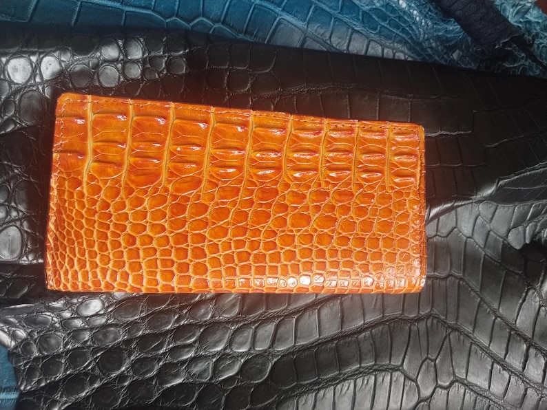 May include: An orange leather wallet with a textured crocodile pattern. The wallet is rectangular and lies on a surface of black and blue leather with a similar embossed design. The wallet is a warm, vibrant color.