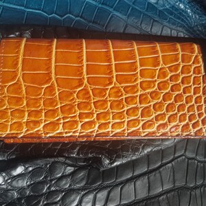 May include: A rectangular, tan-colored wallet with a crocodile skin pattern. The wallet is closed and appears to be made of leather. The background features textured leather in shades of black and blue.
