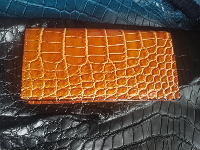 May include: A rectangular, tan-colored wallet with a crocodile skin pattern. The wallet is closed and appears to be made of leather. The background features textured leather in shades of black and blue.