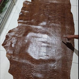May include: A large, irregular piece of brown leather featuring a glossy, embossed crocodile skin texture. The surface displays a repeating pattern of small, rectangular scales with a reflective finish, covering the entire hide from edge to edge.
