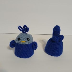 May include: Two handmade crochet items. One is a blue and gray creature with black eyes and a yellow beak. The other is a blue bell-shaped item with a rounded top and two smaller spheres. Both items are on a white surface.