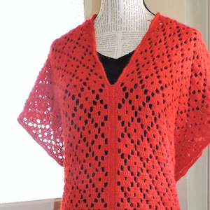 May include: A vibrant red, open-weave crochet garment with a V-neck and short sleeves. The long, flowing design features a central seam and a scalloped hem. The garment is displayed on a mannequin.