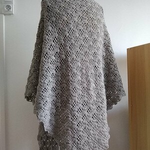 Puede incluir: Un chal de ganchillo gris con un delicado patr&oacute;n de encaje. El chal est&aacute; drapeado sobre un maniqu&iacute; y se muestra contra una pared blanca.