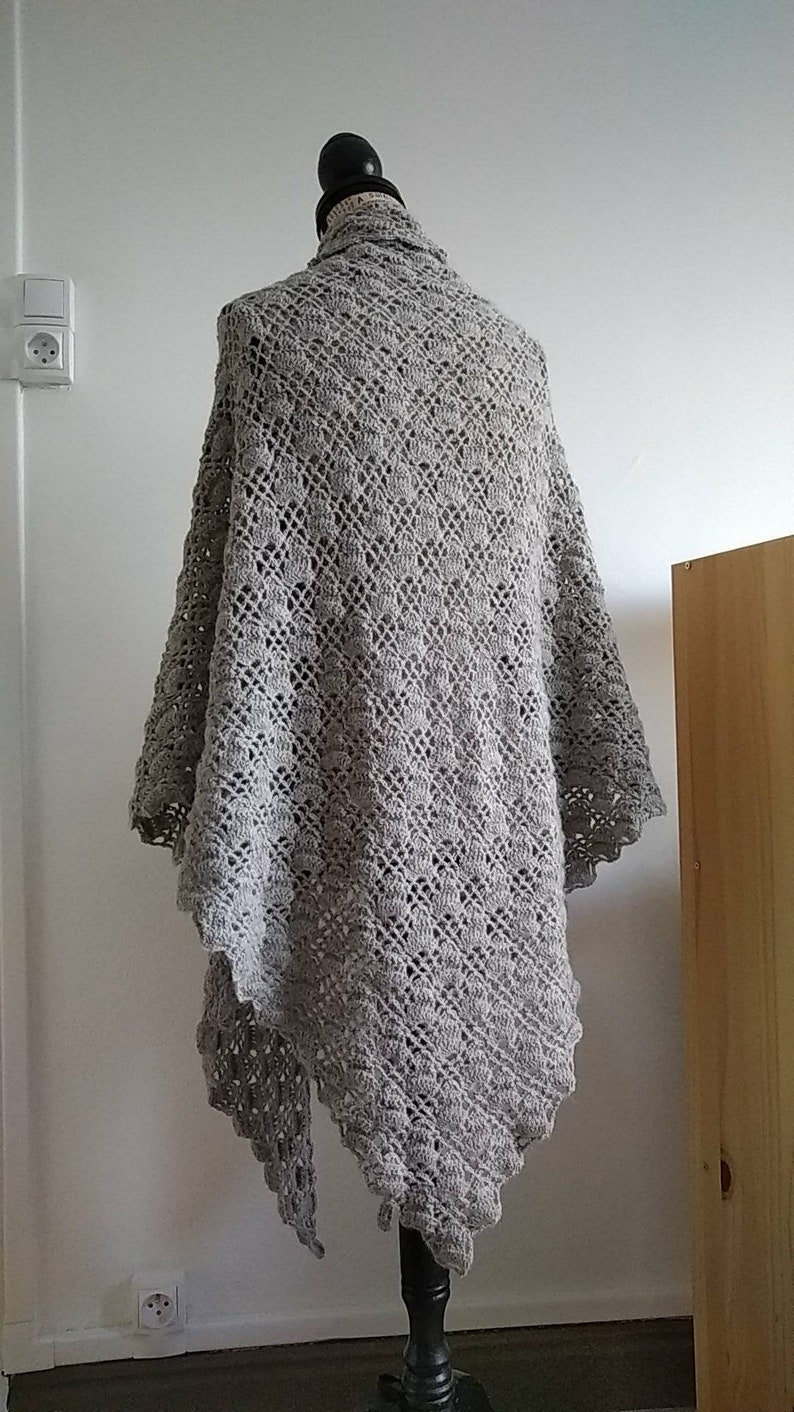 Puede incluir: Un chal de ganchillo gris con un delicado patr&oacute;n de encaje. El chal est&aacute; drapeado sobre un maniqu&iacute; y se muestra contra una pared blanca.