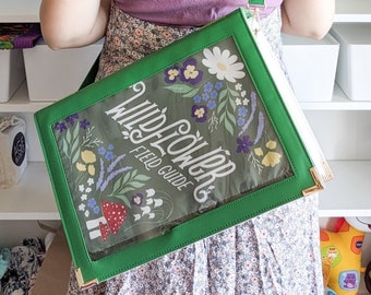 May include: A green and white clear vinyl tote bag with a gold chain strap. The bag has a floral design and the text "Wildflowers Field Guide".