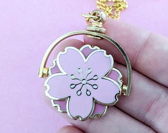 May include: A gold chain necklace with a pink and white cherry blossom charm that spins. The charm is attached to a gold ring.