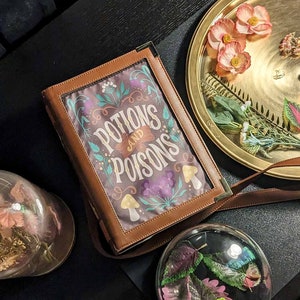 Könnte beinhalten: Eine braune Leder-Buchentasche mit einem floralen Design und dem Text "Potions and Poisons" auf der Vorderseite. Die Tasche liegt auf einer schwarzen Oberfläche mit einer Glaskuppel, die getrocknete Blumen enthält, und einem goldenen Tablett mit getrockneten Blumen.