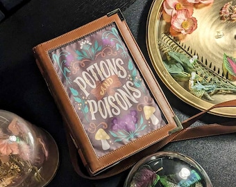 May include: A brown leather book bag with a floral design and the text "Potions and Poisons" on the front. The bag is sitting on a black surface with a glass dome containing dried flowers and a gold tray with dried flowers.