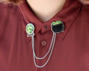 May include: Two silver chains with a green potion bottle and a black cauldron charm attached. The charms are worn as a collar pin on a maroon shirt.