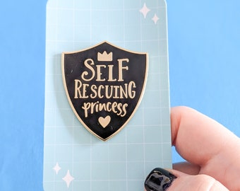 May include: Black enamel pin shaped like a shield with a gold crown, the words "SELF RESCUING princess" and a small gold heart.