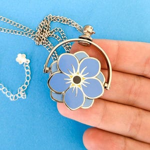 Puede incluir: Collar plateado con un colgante de flor azul. El colgante presenta una flor de esmalte azul claro con un centro amarillo y una cadena plateada. El colgante de la flor está en un marco circular.