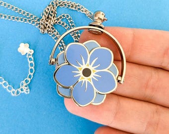 May include: A silver-toned necklace with a blue flower pendant. The pendant features a light blue enamel flower with a yellow center and a silver chain. The flower pendant is in a circular frame.