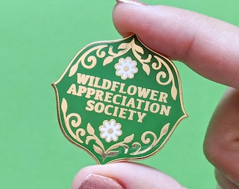 May include: Green enamel pin with gold trim and white flowers. The pin reads "Wildflower Appreciation Society".